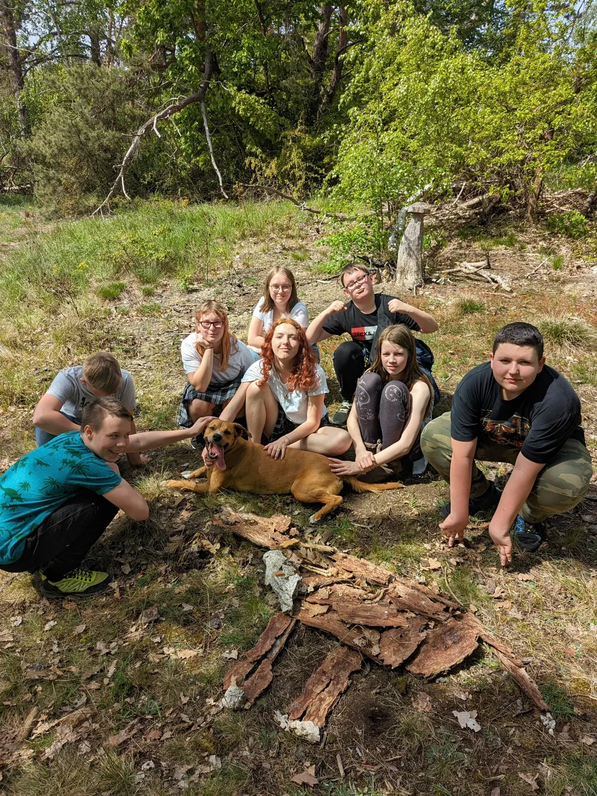 Kinder und Jugendliche hocken mit einem Hund im Wald zusammen bei einem Naturangebot